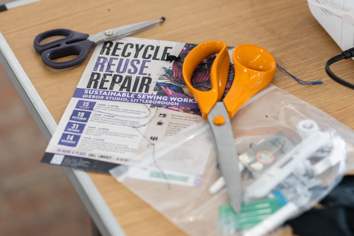 A close-up of a sewing and recycling table scene featuring a pair of orange-handled scissors resting on a 'Recycle Reuse Repair' promo flyer, alongside other sewing tools and a plastic bag of supplies on a wooden desk.