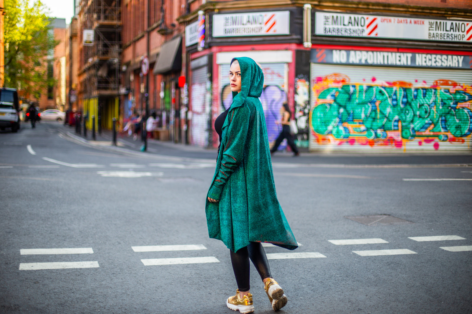 Model Amy wears the Miho cardigan with black wet look leggings, sheer top and gold trainers. She is stood in a road in Manchester in front of some graffiti shutters