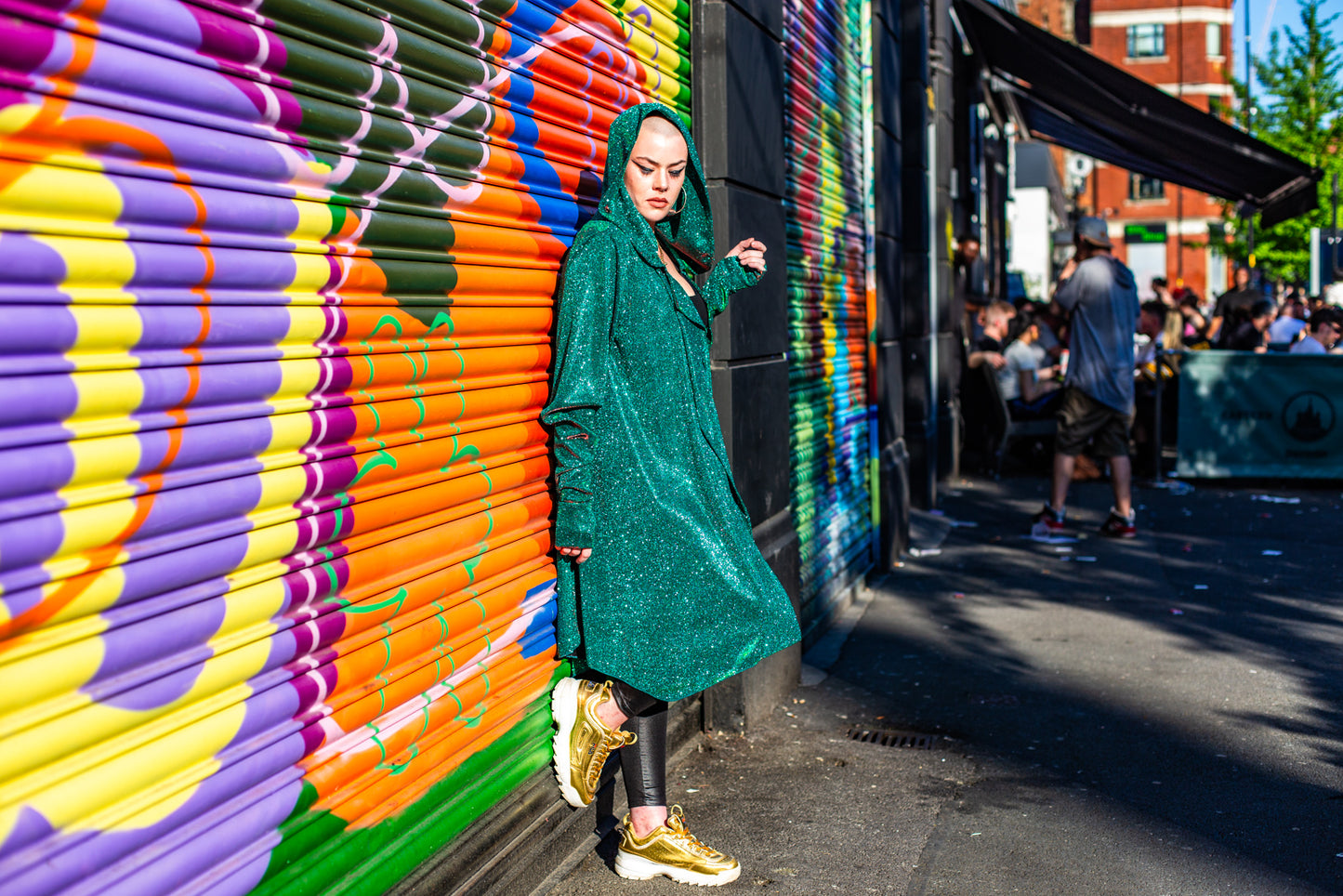 Model Amy wears the Miho cardigan with black wet look leggings, sheer top and gold trainers. She is stood in Manchester in front of some graffiti shutters and bars behind her