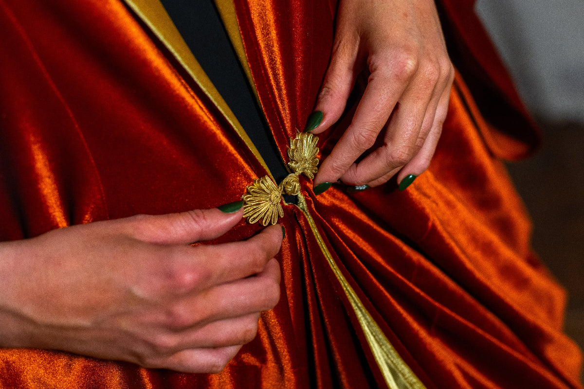 Close-up of a person's hands with green nail polish fastening a gold decorative clasp onto a draped orange-red VF Ophelia with a gold trim.