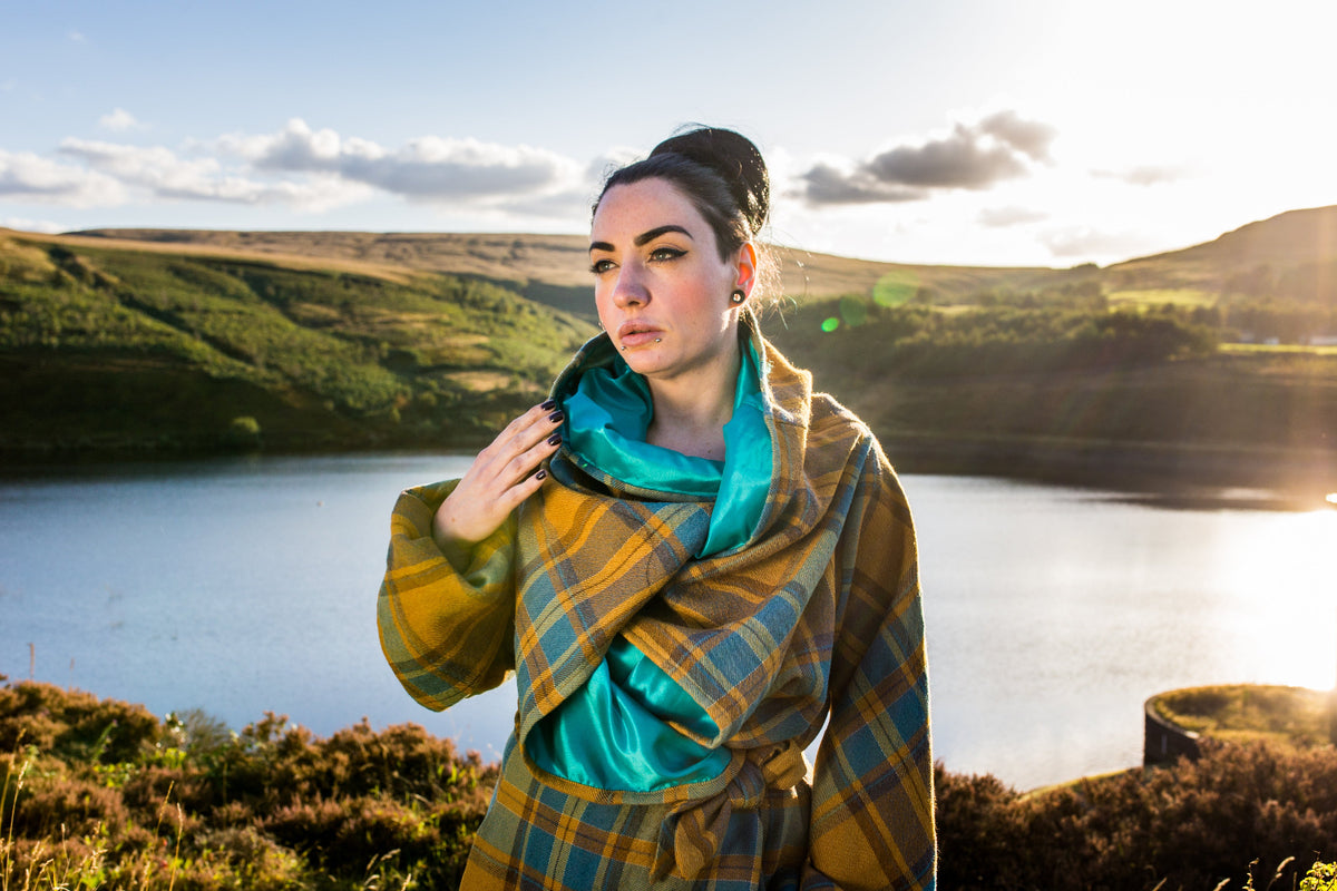 A woman standing outdoors by a lake with hills in the background, wearing a yellow and blue plaid VF Kira trench coat with a bright turquoise lining.
