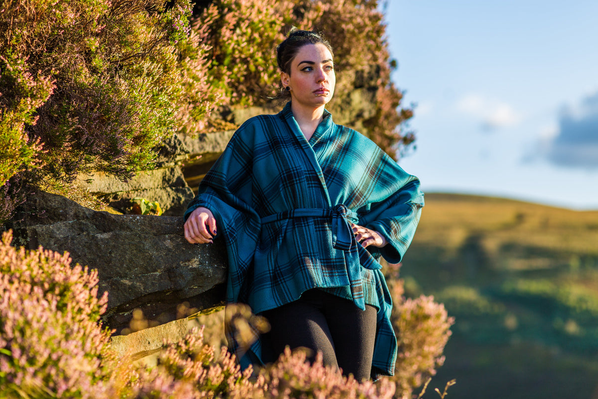 A woman wearing a teal and black plaid VF Orla coat leans against a stone wall outdoors, with heather and a hilly landscape in the background.
