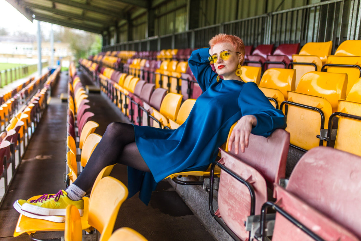 A woman with short pink hair and yellow sunglasses sits in a stadium seating area, wearing a teal VF Maya dress, black tights, and yellow sneakers. The background features rows of yellow and maroon stadium seats under a covered structure.