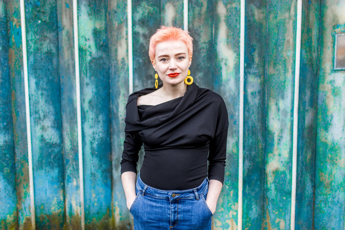 A woman with short pink hair stands in front of a textured teal metal wall, wearing a black draped long-sleeve top, blue jeans, and large yellow hoop earrings.