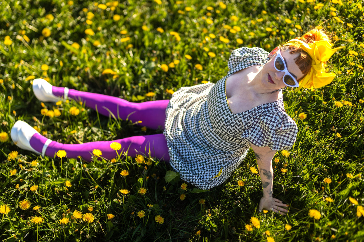 Lindsey the company owner sits in a field of dandelions, wearing a black and white houndstooth dress and sunshine yellow headwrap