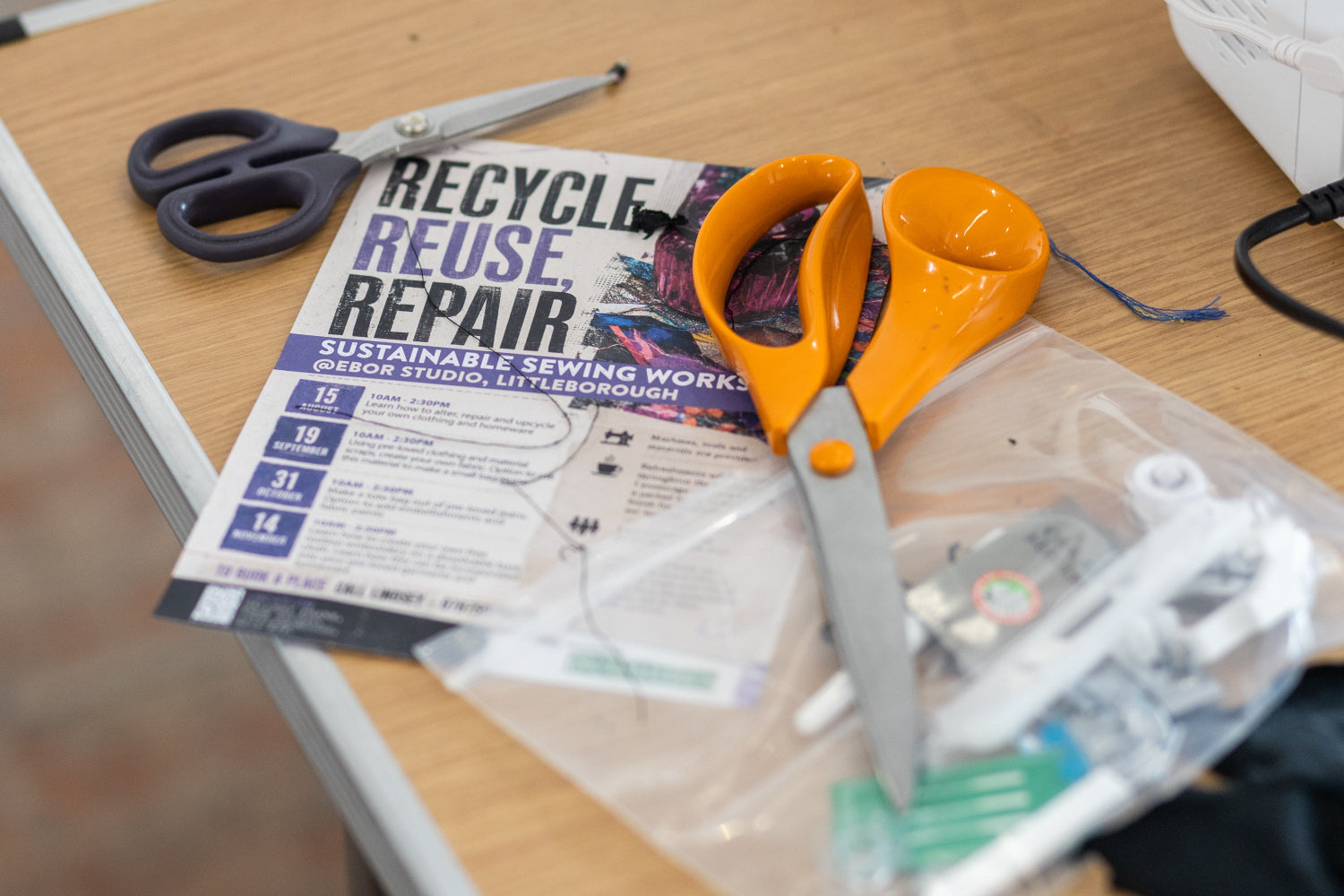 A close-up of a sewing and recycling table scene featuring a pair of orange-handled scissors resting on a 'Recycle Reuse Repair' promo flyer, alongside other sewing tools and a plastic bag of supplies on a wooden desk.