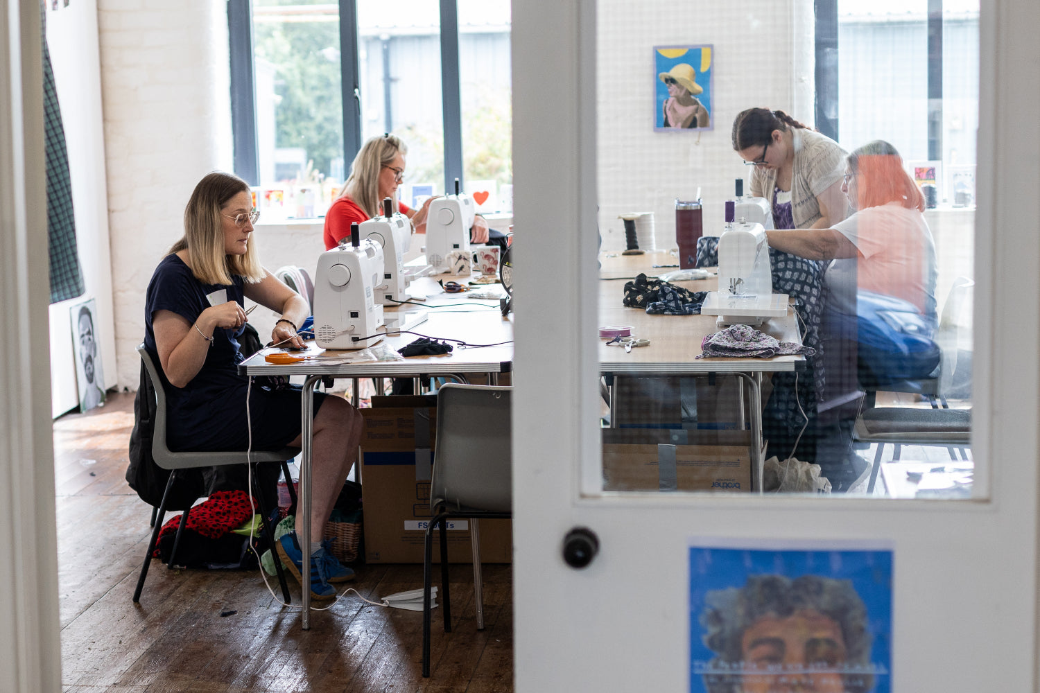 Gallery Fank at Ebor Studio with large windows and a glass door in the foreground, showing several women working at sewing tables with sewing machines and fabric. Because people and a real environment are clearly present, this is Lifestyle rather than Product/Packaging.